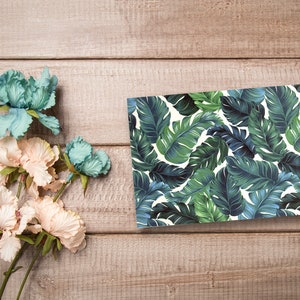May include: A flat card with a tropical leaf pattern in shades of green and blue on a white background. The card is on a wooden surface with a bouquet of light pink and blue flowers.