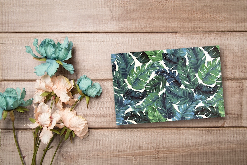May include: A flat card with a tropical leaf pattern in shades of green and blue on a white background. The card is on a wooden surface with a bouquet of light pink and blue flowers.