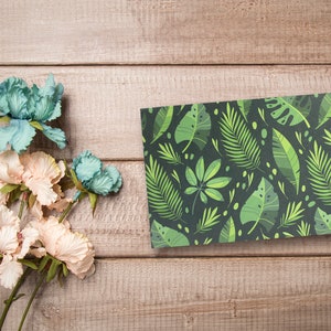 May include: A flat card with a green and black tropical leaf pattern. The card is on a light brown wooden surface with a bouquet of light pink and blue flowers.