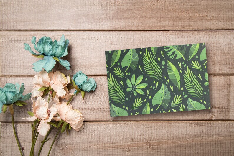 May include: A flat card with a green and black tropical leaf pattern. The card is on a light brown wooden surface with a bouquet of light pink and blue flowers.