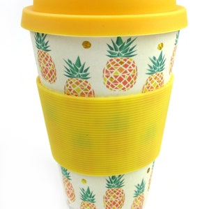 May include: A yellow and white reusable coffee cup with a pineapple pattern. The cup has a yellow lid and a yellow silicone band around the middle.