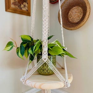 May include: A macrame plant hanger with a wooden shelf and a green potted plant. The hanger is made of white cotton cord and has a geometric pattern. The shelf is round and has a natural wood finish.
