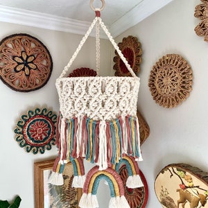 May include: A white macrame hanging mobile with rainbow-colored yarn tassels and a white crocheted basket. The mobile is hanging from a wooden ring and is decorated with woven wall decor.