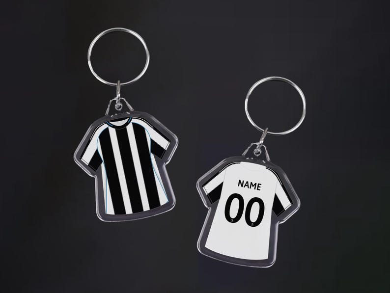 May include: Two clear acrylic keychains shaped like soccer jerseys. One features black and white stripes, the other is white with black accents, the word "NAME" and the number "00". Each keychain has a silver keyring.