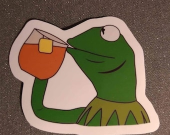 May include: A green frog wearing a yellow and green collar is drinking from a glass of orange liquid with a tea bag inside. The frog has a white eye and is looking to the right.