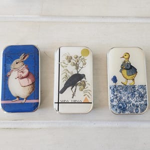 May include: Three rectangular metal tins with rounded corners. The left tin is blue with a rabbit in a pink coat. The center tin is white with a black bird on a branch, a yellow circle, and the text SHINY THINGS. The right tin features a yellow duckling.