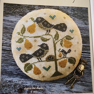 May include: A round, beige cross-stitch design featuring a tree with pears and birds. The birds are black and the pears are yellow. Green leaves and teal accents complete the design. The text "Scattered Seed Samplers © 2022" is at the bottom.