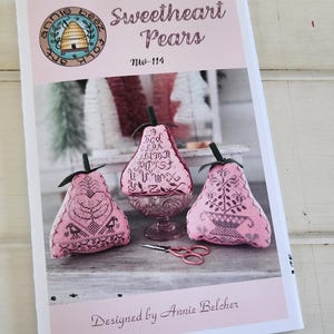 May include: A pink and white cross stitch pattern booklet for a craft project called "Sweetheart Pears" by Annie Bee Folk Art. The booklet cover features a beehive logo and three pink pear-shaped pincushions.
