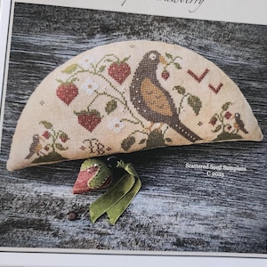 Può includere: Un ricamo a punto croce semicircolare su tessuto beige con un uccello marrone, fragole rosse e foglie verdi. Sotto, un piccolo puntaspilli a forma di fragola con un nastro verde. Il testo recita Scattered Seed Samplers C 2025.