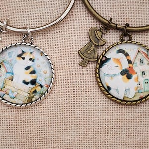 May include: Two round pendant bracelets with cat illustrations. One pendant has a silver finish and a cat sitting on a wooden structure. The other pendant has a gold finish and a cat in front of a house. Each bracelet has a charm.