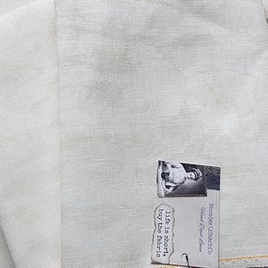 May include: Two folded pieces of off-white linen fabric. A small tag with a vintage image and text is attached. The tag reads "Life is short, buy the fabric."