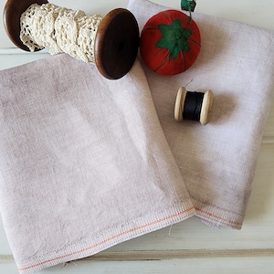 May include: Two pieces of light gray linen fabric with orange stitching. A spool of white lace wound around a wooden spool, a red pincushion shaped like a tomato, and a spool of black thread wound around a wooden spool are on top of the fabric.