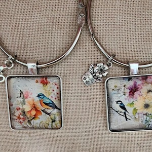 Può includere: Due bracciali rigidi in metallo color argento, ciascuno con un ciondolo quadrato con un'immagine stampata di uccelli e fiori. Un ciondolo mostra un uccello blu con accenti arancioni, l'altro un uccello bianco e nero. Ogni bracciale ha un piccolo ciondolo.