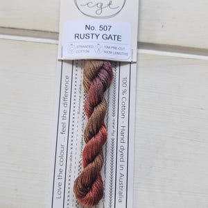 May include: A skein of hand-dyed stranded cotton embroidery thread in the color Rusty Gate, No. 507. The thread features a variegated blend of brown, copper, and deep red tones, mounted on a white card with 10M pre-cut 50cm lengths printed on it.