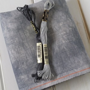 Puede incluir: Un trozo de tela de lino gris con dos carretes de hilo de bordar, uno negro y otro gris. Los carretes tienen etiquetas con números y códigos de barras.