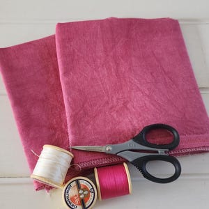 May include: Two pieces of pink fabric, a spool of white thread, a spool of pink thread, and a pair of silver scissors on a white wooden surface.