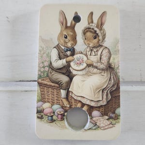 May include: A rectangular decorative plaque with a vintage illustration of two rabbit characters. One rabbit wears a waistcoat and monocle, the other a bonnet. They are seated on a wicker basket, holding an embroidered hoop. Pastel eggs and thread spools are also visible.