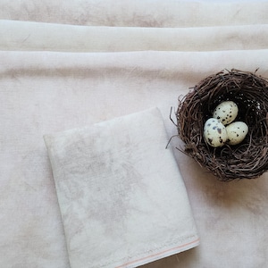 May include: A set of two off-white linen fabric squares with a subtle speckled pattern. One square is folded in thirds and the other is flat. A small woven bird's nest with three speckled quail eggs sits on the fabric.