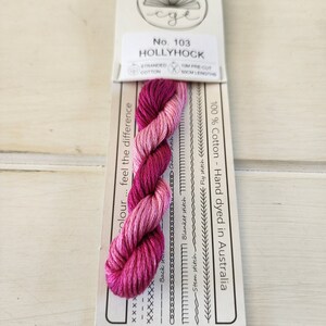 Puede incluir: Un ovillo de hilo de bordar de algodón Hollyhock, teñido a mano en Australia. El hilo presenta un degradado de tonos rosa y magenta. El embalaje incluye el texto "No. 103 HOLLYHOCK" y ejemplos de puntadas.