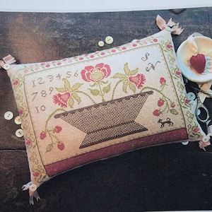 May include: A rectangular, embroidered pillow with a floral design and the numbers 1-10. The pillow features a basket with flowers and a small dog. Pink ribbons and buttons adorn the corners. A small bowl with fruit and scissors are nearby.