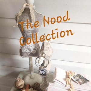 May include: Two miniature dress forms, one grey with white floral print and one beige, stand behind a small wooden basket filled with white buttons, a pincushion, and a stack of folded light pink fabric tied with lace and a small rectangular tag.