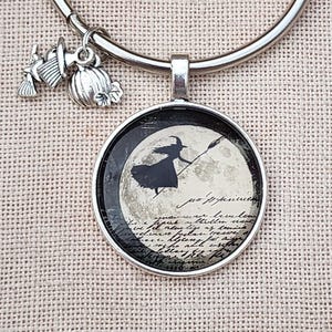 Può includere: Un bracciale rigido color argento con un ciondolo rotondo raffigurante la sagoma nera di una strega che vola su una scopa davanti a una luna piena con testo in corsivo. Due piccoli ciondoli argentati, una scopa e una zucca, sono appesi.