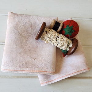 Op de afbeelding: Twee roze linnen theedoeken met oranje stiksels, een houten spoel met witte kant en een rood naaldenkussen met groene bladeren.