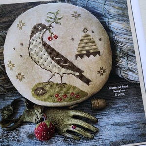 Pode incluir: Um sampler redondo de ponto cruz com fundo bege que apresenta um pássaro marrom e branco com bagas vermelhas, uma colmeia e um desenho floral verde e marrom. O texto "Scattered Seed Samplers C 2024" está impresso abaixo do sampler.