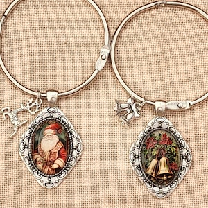May include: Two silver-toned metal circular rings, each featuring a diamond-shaped pendant. One pendant displays a vintage-style illustration of Santa Claus, while the other shows two golden bells with holly. Each ring includes a small dangling charm.