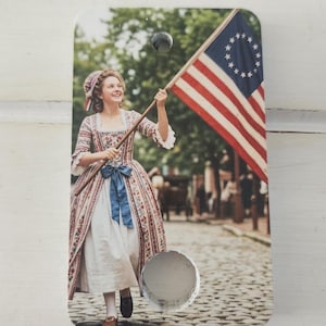 Könnte beinhalten: Rechteckige Holzplakette mit einem historischen Bild einer Frau in Kolonialkleidung, die die amerikanische Flagge hält. Die Flagge hat rote und weiße Streifen und einen Sternenkreis. Die Frau lächelt und geht auf einer Kopfsteinpflasterstraße.