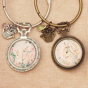 May include: Two charm bracelets with pocket watch pendants. One bracelet is silver-toned, the other is bronze. Each pendant features a watercolour-style bunny illustration. The silver bracelet has a floral charm, the bronze has a watering can charm.