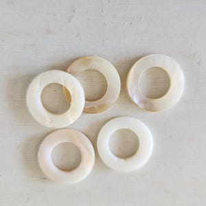 May include: Five circular rings made of iridescent mother-of-pearl material. Each ring features a smooth, polished surface with natural variations in colour, ranging from creamy white to soft beige tones, showcasing a distinct pearlescent lustre.