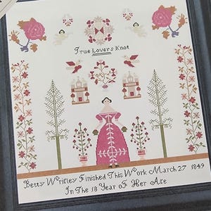 May include: Framed cross-stitch sampler with a black frame. The design features a central figure in a pink dress, surrounded by floral and architectural motifs in pink, red, and green. Text includes "True Lovers Knot" and "Betty Wrigley Finished This Work March 27 1849".