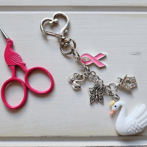 May include: A pair of pink embroidery scissors with a bird design and a silver keychain with charms. The charms include a pink ribbon, a butterfly, a swan, and a small birdhouse.