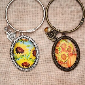 Op de afbeelding: Twee armbanden met ovale hangers met zonnebloem- en pompoenontwerpen. Eén armband is zilverkleurig met een slakkenbedel en eikelbedel. De andere armband is bronskleurig met een slakkenbedel en eikelbedel.