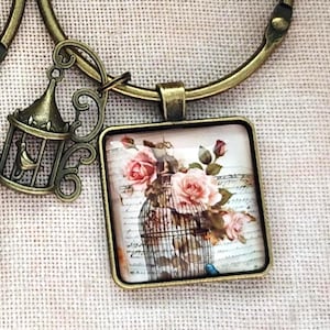 Puede incluir: Un collar de tono bronce con un colgante cuadrado que presenta un cabujón de cristal con una ilustración de rosas rosadas en una jaula de pájaros sobre un fondo de escritura, acompañado por un pequeño dije de jaula en un eslabón circular.