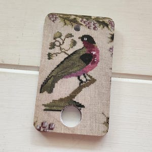Puede incluir: Objeto rectangular beige con un diseño de pájaro bordado a punto de cruz. El pájaro tiene cabeza negra, pecho rosa y alas verdes, posado en una rama. El objeto tiene un agujero circular.
