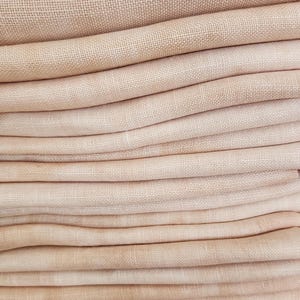 May include: A stack of folded, light pink linen fabric. The fabric has a subtle woven texture and a soft, muted color palette. The folds create a layered effect, showcasing the fabric's drape and volume. The overall aesthetic is simple and elegant.