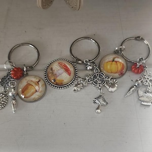 May include: Three circular keychains featuring autumn-themed cabochons. The first shows an acorn, the second a mushroom, and the third a pumpkin. Each keychain includes silver-toned metal charms shaped like hedgehogs, umbrellas, birds, and Celtic knots.