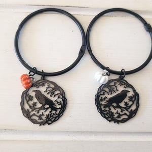 May include: Two black circular bangle bracelets, each featuring a circular charm with a cutout design of a crow perched on a tree branch. One bracelet includes a small orange pumpkin charm, while the other features a small white pumpkin charm.