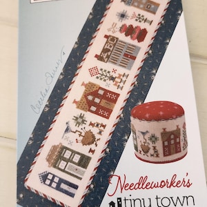 Può includere: Un modello a punto croce e un cuscinetto per spilli sono in mostra. Il modello presenta case ricamate in vari colori, con il testo "Needleworker's Tiny Town". Il cuscinetto per spilli è rotondo con la parte superiore rossa e un disegno di casa ricamata.