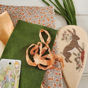 Può includere: Una collezione di materiali per l'artigianato tra cui tessuto, feltro e nastro. È presente anche un oggetto a forma di carota con un disegno di coniglio. I tessuti presentano motivi floreali e colori a tinta unita. La tavolozza dei colori è terrosa, con verdi, arancioni e creme.