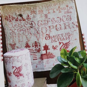May include: A white linen sampler with red embroidery featuring the words "To God Give Grace" and "Sweet to Be Loved". The sampler also includes a bird, a swan, and a tree. A small white cylinder with red embroidery is in the foreground.