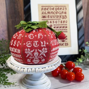 Op de afbeelding: Een rode, ronde, geborduurde ornament met witte kruissteekpatronen en letters, inclusief de tekst "C+H". Het ornament heeft een groen lint en staat op een witte, kralen taartstandaard. Een ingelijste borduurwerk staat op de achtergrond.