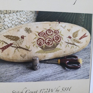 May include: Oval-shaped pin cushion with a cross-stitched design featuring birds, flowers, and a basket. The cushion is beige with brown and red embroidery. A silver thimble and scissors are also visible.