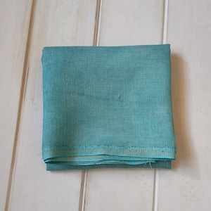 以下が含まれることがあります： 微妙なテクスチャのターコイズブルーの布を折りたたんだもの。布の端には金色の糸が走っています。