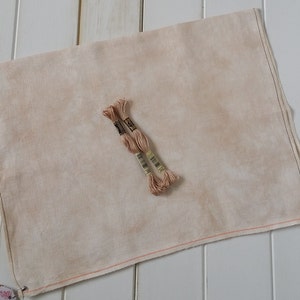 May include: Two skeins of light brown embroidery floss on a beige linen fabric with a red stitched border.