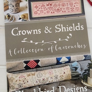 May include: A book cover titled "Crowns & Shields: A Collection of Cartouches" by Blackbird Designs. The cover features cross-stitch samplers and a framed design. The color palette includes shades of gray, pink, and cream.