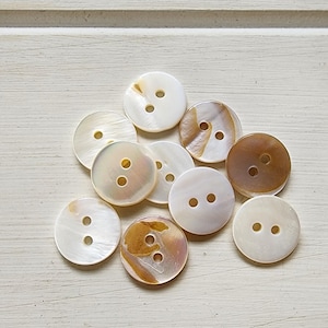 May include: A selection of round buttons with two holes, in shades of white, cream, and brown. The buttons have a pearlescent finish, some with a marbled appearance. They are arranged on a light-coloured, textured surface.