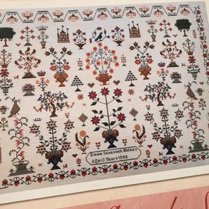 Könnte beinhalten: Ein rechteckiges Stickmustertuch mit komplexer Kreuzstichstickerei auf hellem Stoff. Das Design umfasst symmetrische Blumenmotive, Bäume, Vögel und architektonische Elemente in Schwarz, Rot und Blau. Der Text lautet Emma Susannah Helman Age 13 Years 1849.